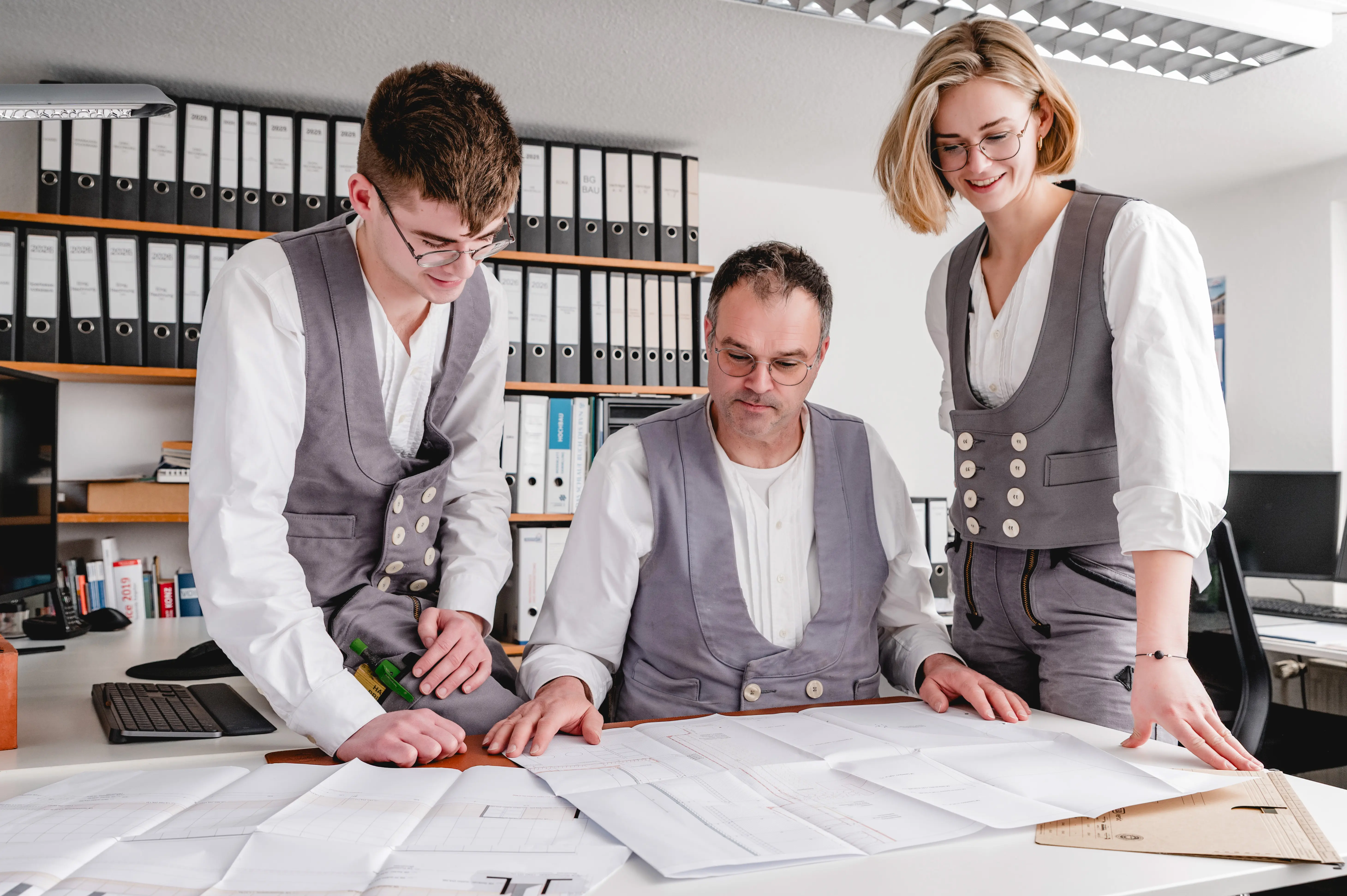 Team Sommerfeld: Erfahrener Bauleiter mit jüngeren Kollegen in traditioneller Arbeitskleidung besprechen detaillierte Baupläne für ein Sanierungsprojekt im Büro.